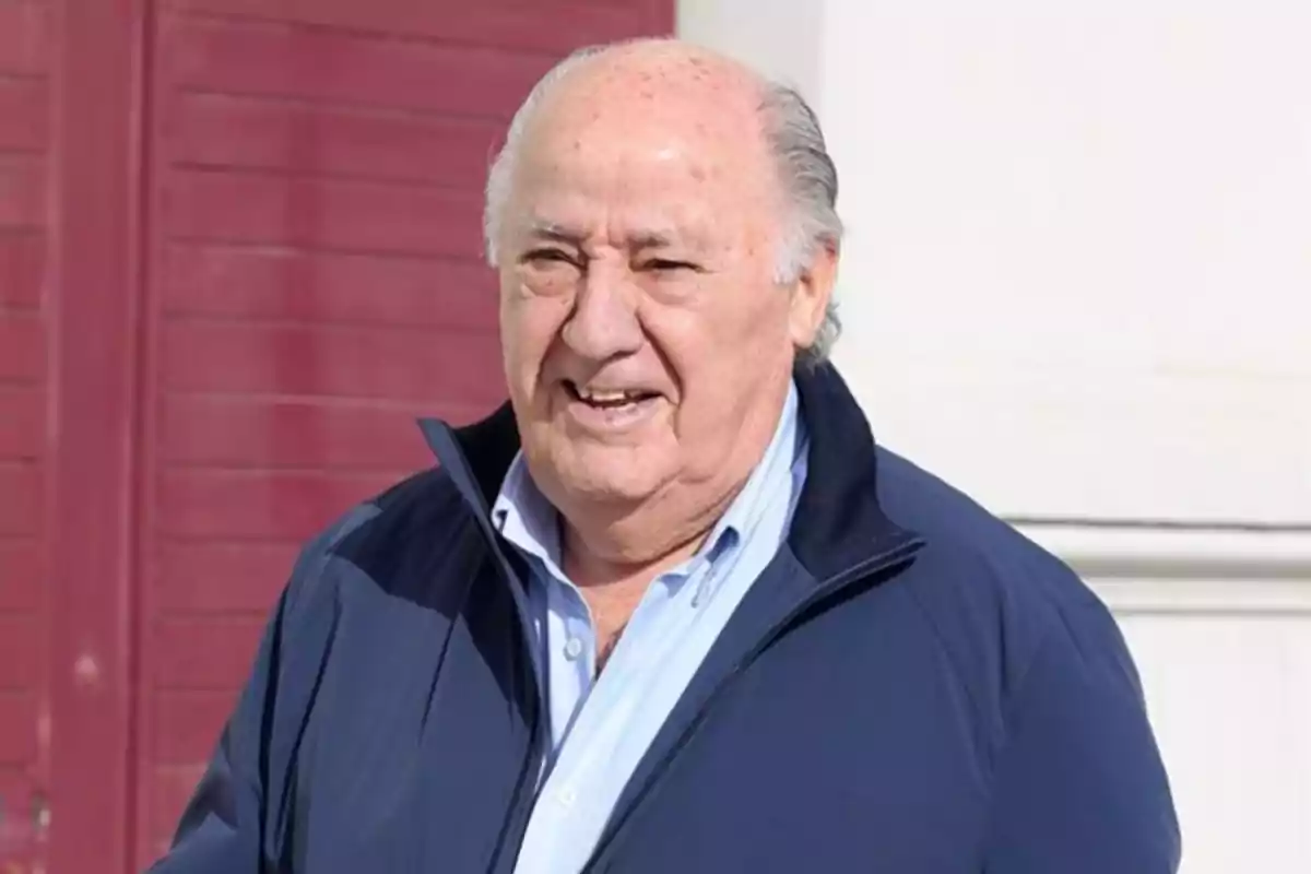 Amancio Ortega con chaqueta azul y camisa clara sonríe mientras está de pie frente a una pared de color rojo oscuro.