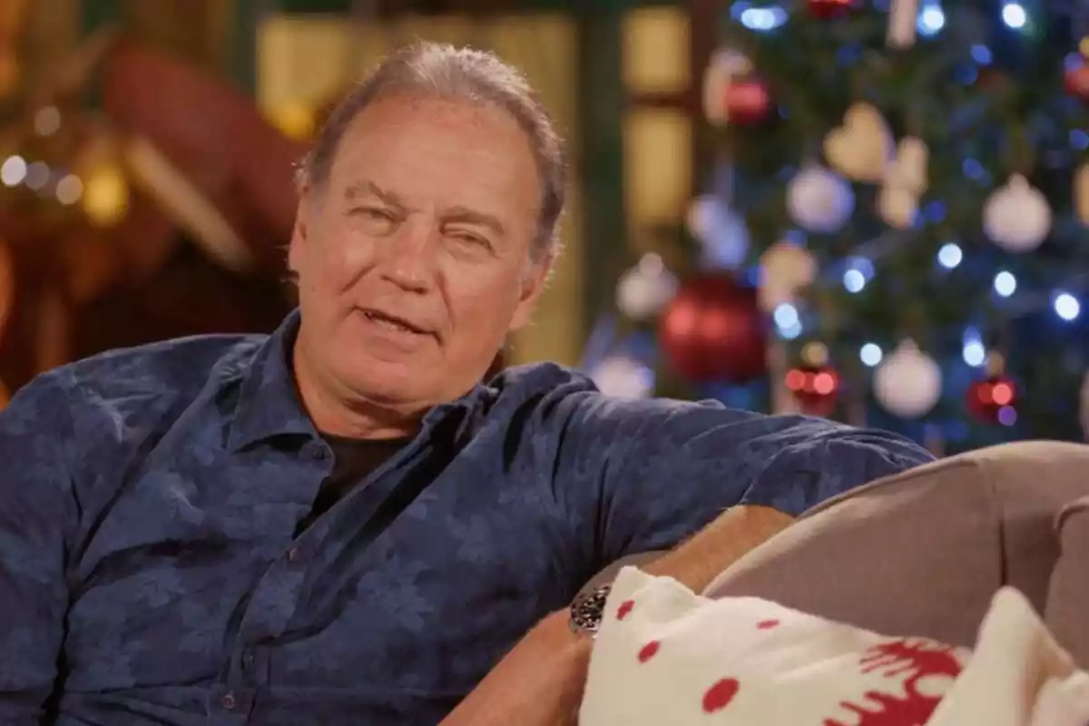 Imagen de Bertín Osborne sentado en el sofá en una entrevista para Telecinco. Lleva una camisa con estampado de flores azul y en el fondo hay un árbol de Navidad con las luces encendidas