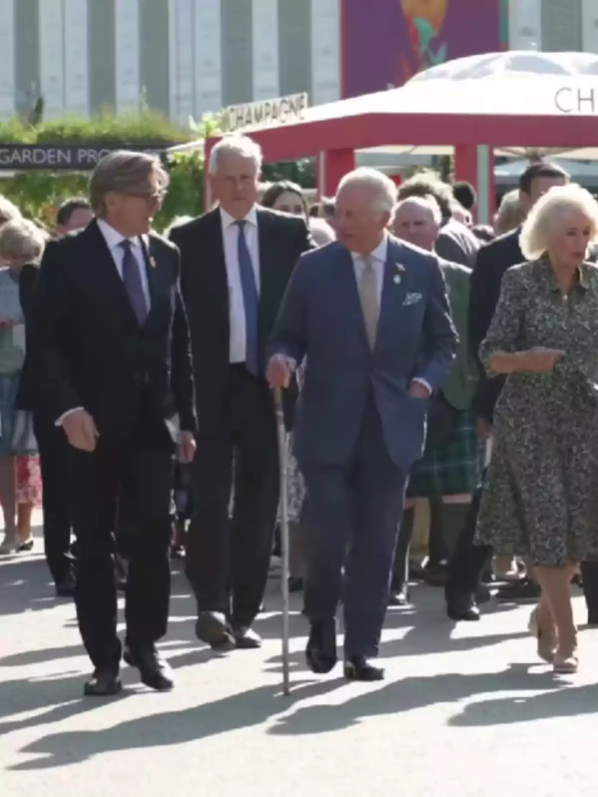 A group of people walking outdoors at an event, with an older man using a cane and a woman in a patterned dress. A group of people walking outdoors at an event, with an older man using a cane and a woman in a patterned dress.