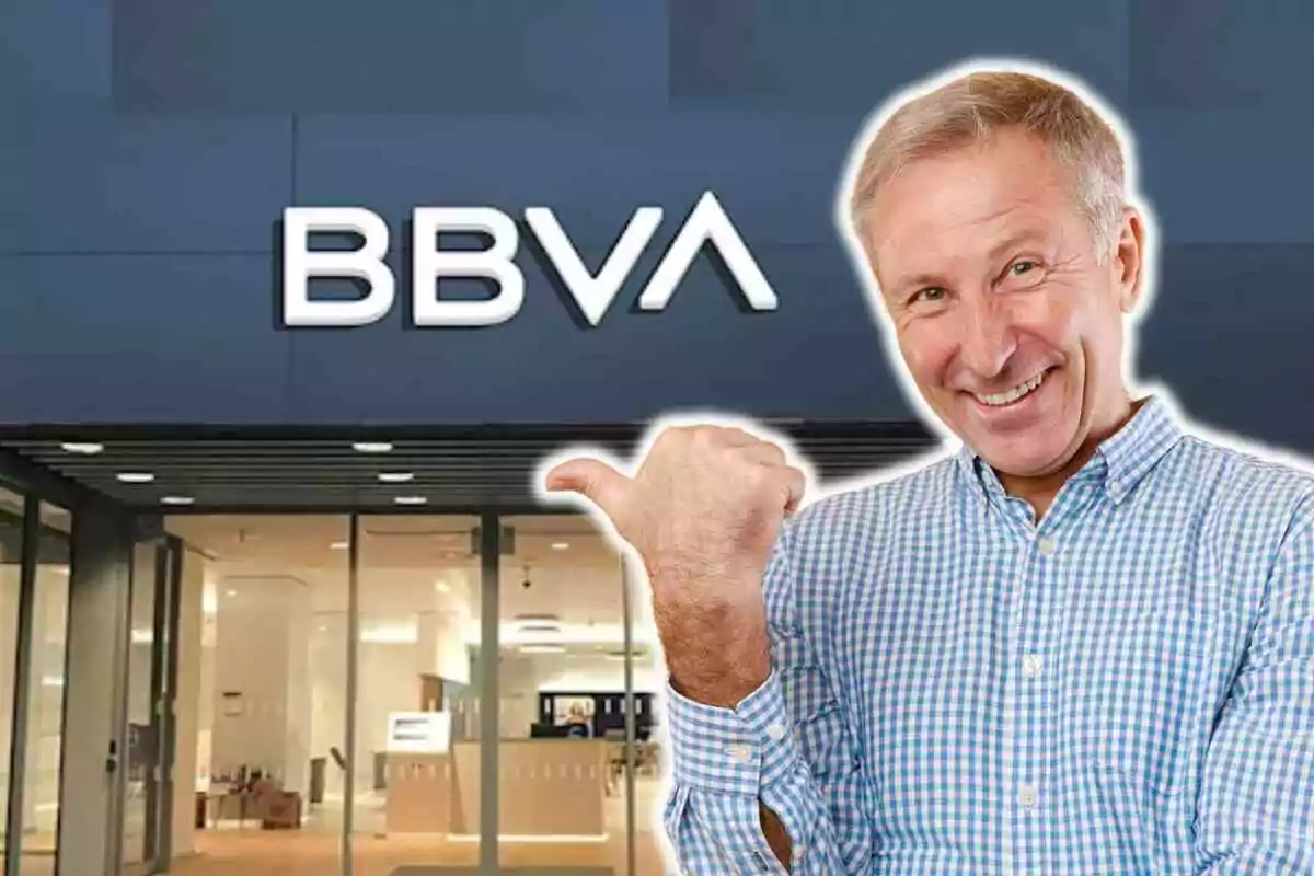 Un hombre sonriente con camisa a cuadros azules y blancos señala con el pulgar hacia una sucursal del banco BBVA. Un hombre sonriente con camisa a cuadros azules y blancos señala con el pulgar hacia una sucursal del banco BBVA.