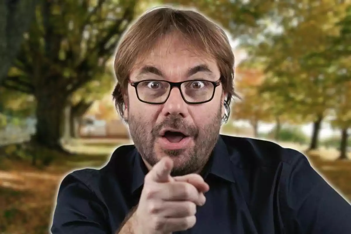 Hombre con gafas y barba señalando con el dedo en un entorno natural otoñal.