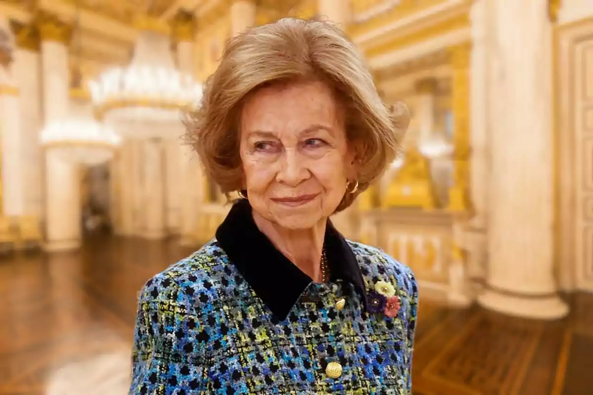 Una mujer mayor con cabello castaño claro y un abrigo de colores en un elegante salón con candelabros y columnas.