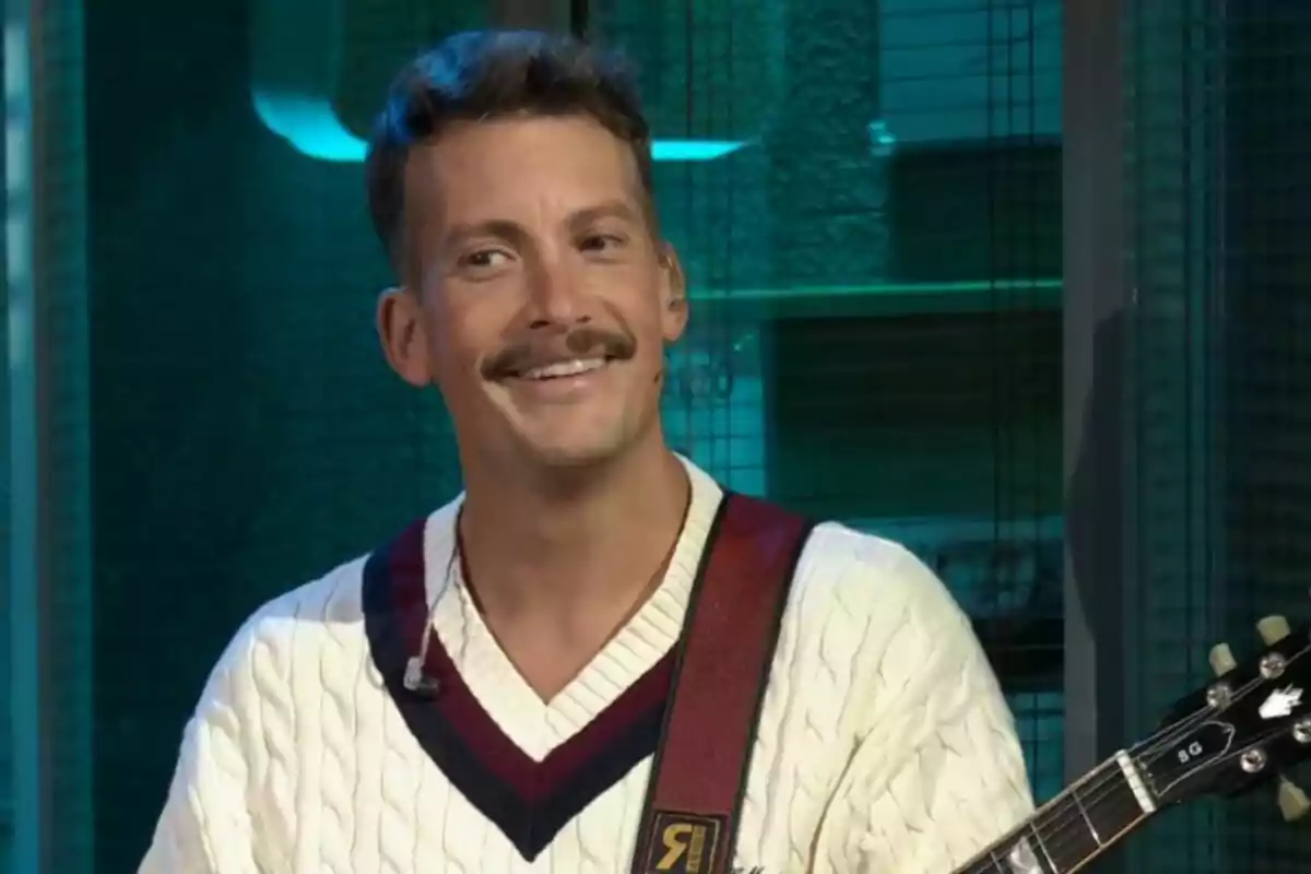 Grison sonriente con bigote y suéter blanco sostiene una guitarra en La Revuelta.