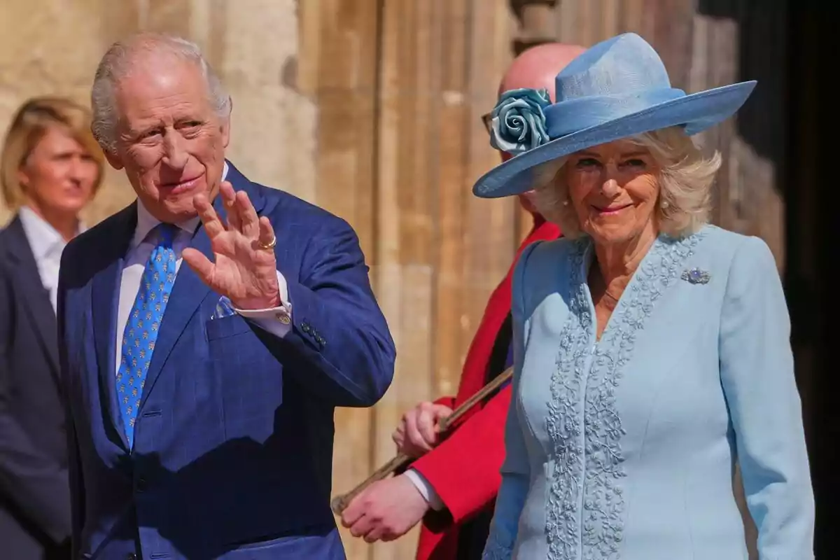 Dues persones grans elegantment vestides, una d'elles amb un vestit blau i corbata a joc, saluden mentre caminen a l'aire lliure, l'altra persona porta un vestit blau clar amb un barret adornat amb una flor.