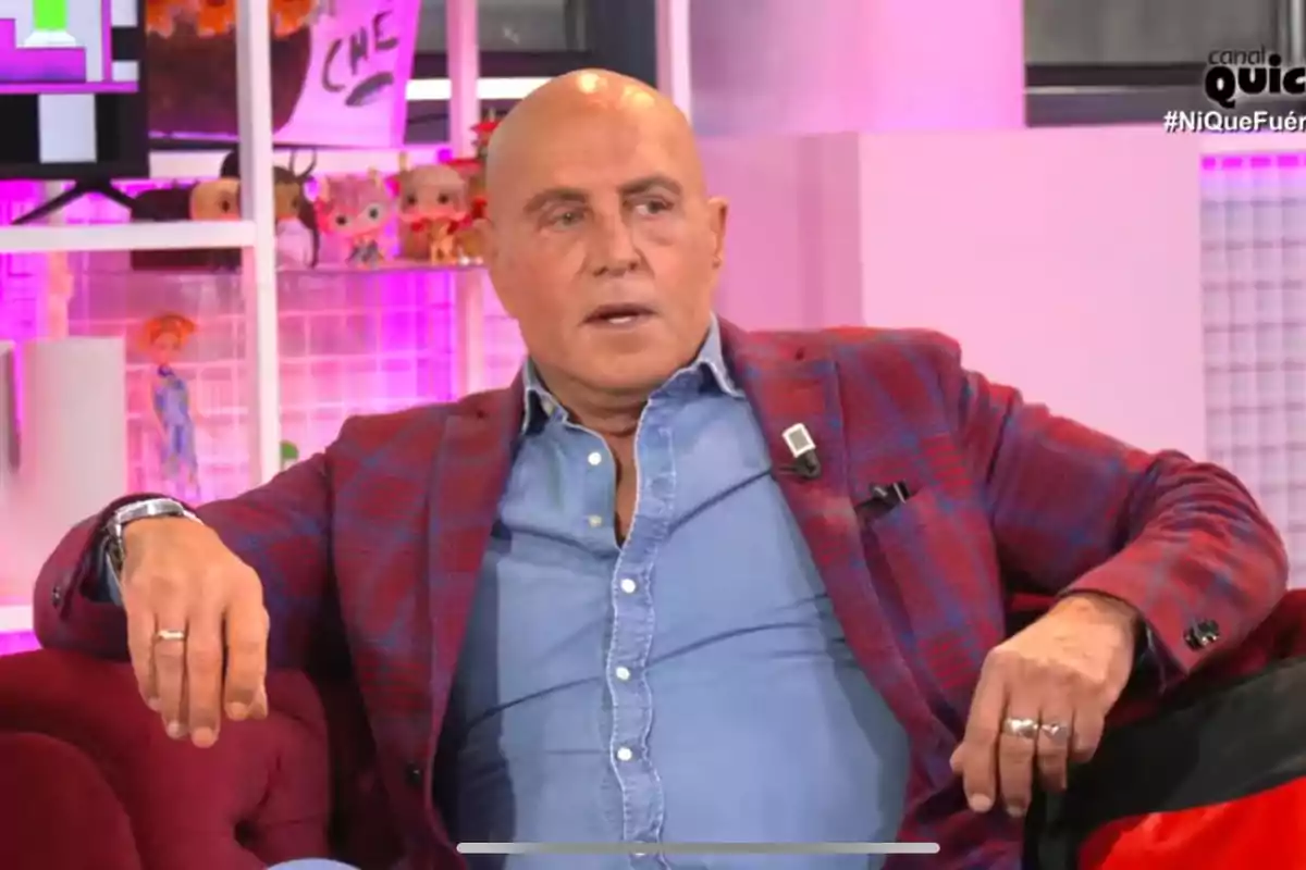 Kiko Matamoros con chaqueta a cuadros y camisa azul está sentado en un sofá en un set de televisión con luces rosadas y juguetes en el fondo.