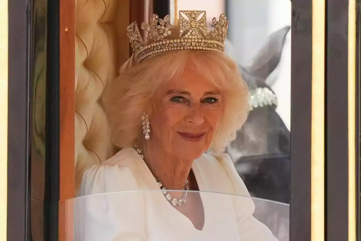 Una dona amb cabell ros i una corona adornada amb joies, vestida de blanc, somriu des de l'interior d'un carruatge.