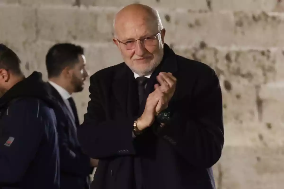 Juan Roig con barba y gafas aplaude mientras viste un abrigo oscuro en un entorno de pared de piedra.