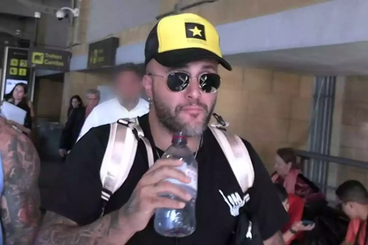 Kiko Rivera serio con una gorra amarilla y negra, gafas de sol y una botella de agua en la mano en el aeropuerto