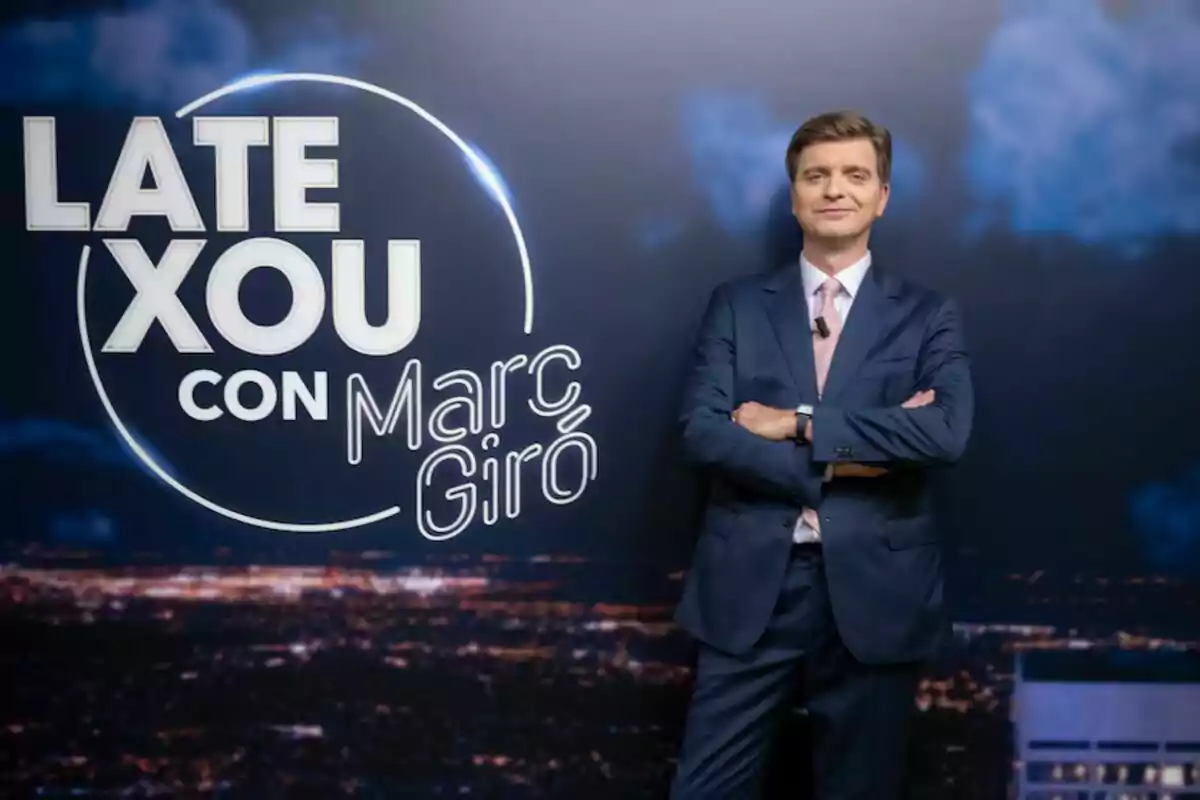 Marc Giró con un traje posa con los brazos cruzados frente a un fondo que publicita su programa, "Late Xou con Marc Giró".