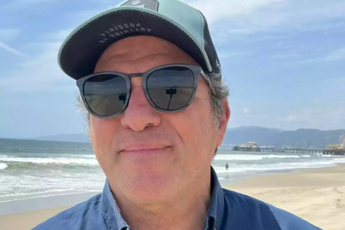 Hombre con gafas de sol y gorra en la playa con el mar y un muelle al fondo.