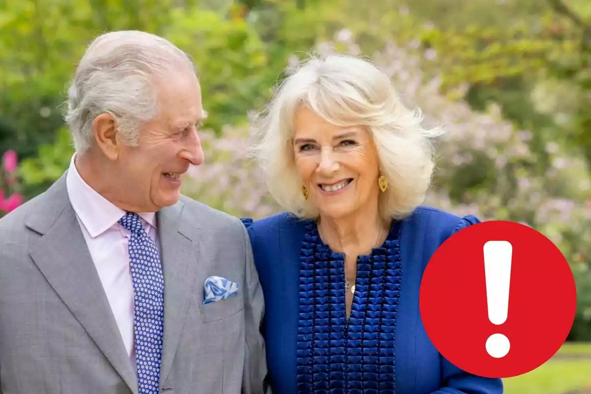 Charles III and Camilla Parker smiling, the man in a grey suit and blue tie, the woman in a blue dress, in an outdoor setting with vegetation in the background and a red exclamation mark icon in the lower right corner. Charles III and Camilla Parker smiling, the man in a grey suit and blue tie, the woman in a blue dress, in an outdoor setting with vegetation in the background and a red exclamation mark icon in the lower right corner.