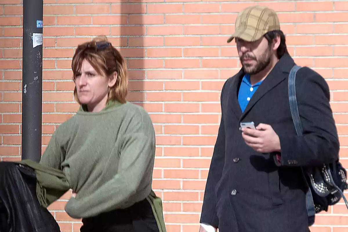 Dos personas caminando por la calle, una mujer con suéter verde y un hombre con gorra y abrigo oscuro, ambos frente a una pared de ladrillos. Dos personas caminando por la calle, una mujer con suéter verde y un hombre con gorra y abrigo oscuro, ambos frente a una pared de ladrillos.