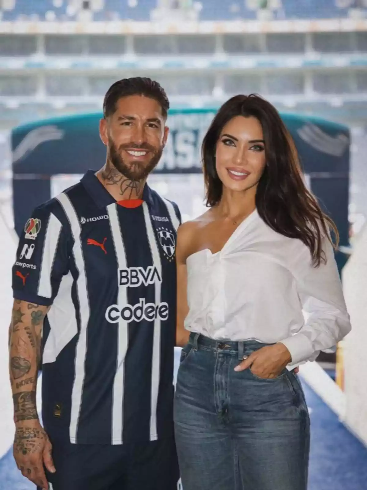 Sergio Ramos in a soccer jersey and Pilar Rubio in a white blouse pose together smiling in a stadium. Sergio Ramos in a soccer jersey and Pilar Rubio in a white blouse pose together smiling in a stadium.
