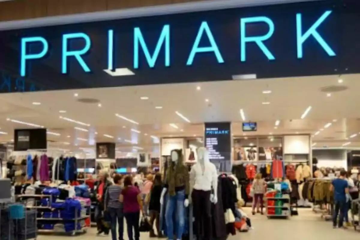 Entrada de una tienda de ropa con el letrero "Primark" iluminado en azul y varias personas comprando en el interior.