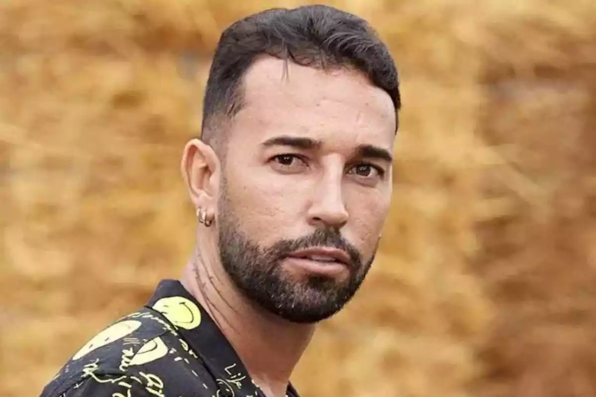 Un hombre con barba y cabello corto mira hacia la cámara con un fondo desenfocado de color marrón.