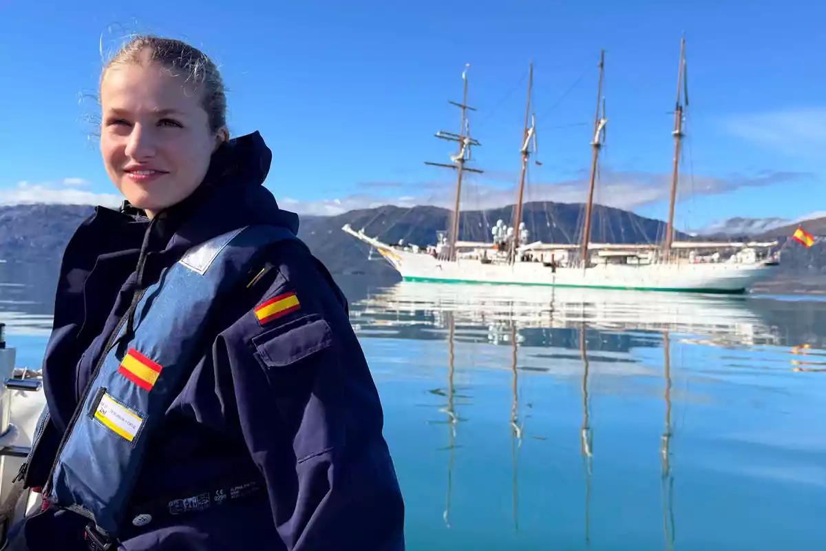 La princesa Leonor amb uniforme blau marí i banderes d'Espanya a l'armilla salvavides està en un vaixell amb un veler de fons en un paisatge muntanyós i cel clar.