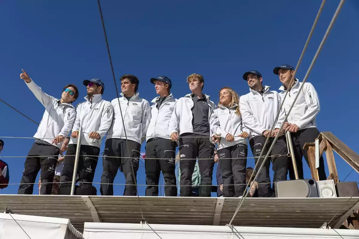 Un grupo de personas vestidas con chaquetas blancas y pantalones oscuros está de pie sobre una estructura al aire libre bajo un cielo azul claro.